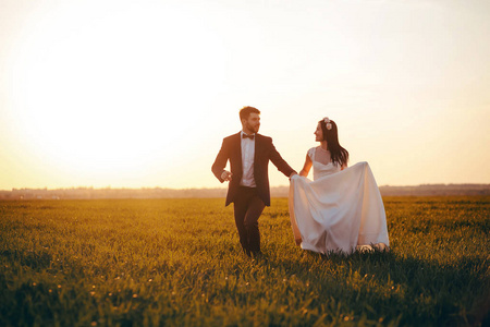 新婚夫妇牵着手跑过田野。 美丽的女孩穿着白色的裙子和时尚的新郎美丽的日落。