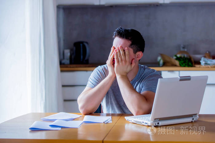年轻疲惫的男人用手抱着头在网上找工作,坐在厨房里拿着笔记本电脑.