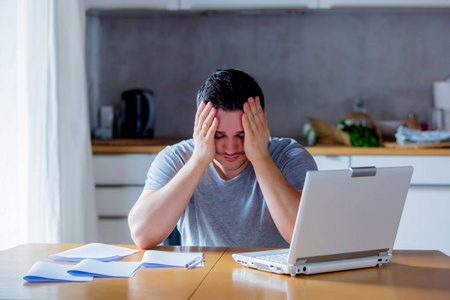 年轻疲惫的男人用手抱着头在网上找工作，坐在厨房里拿着笔记本电脑。