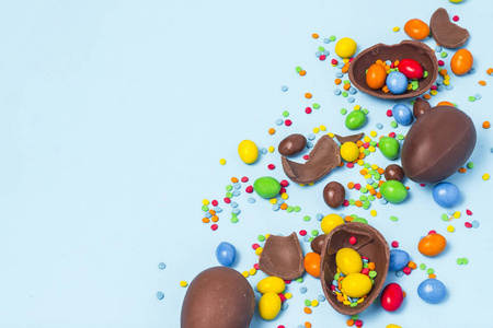 破碎的和整个巧克力复活节鸡蛋，五颜六色的糖果在蓝色背景。 庆祝复活节装饰的概念，寻找复活节兔子的糖果。 平躺顶部视图。 复制空间
