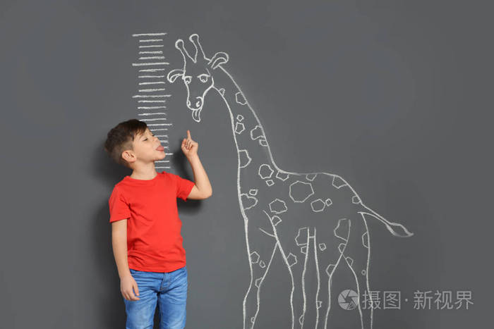 可爱的小孩子在灰色背景下测量身高一边玩粉笔画的长颈鹿