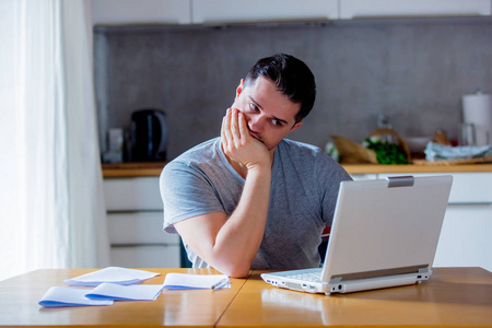 疲惫的年轻人用手摸着嘴在网上找工作，而坐在厨房里用笔记本电脑。