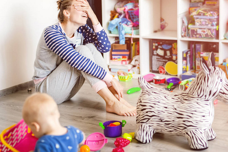 厌倦了日常的家庭母亲坐在地板上, 双手蒙着脸。孩子在凌乱的房间里玩。有秩序的玩具和混乱。快乐的育儿