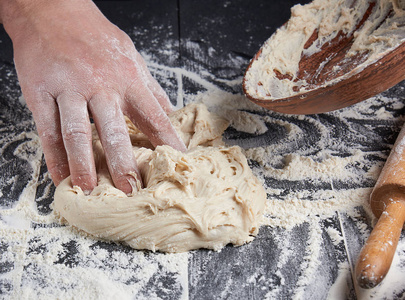 s hands knead white wheat flour dough on black wooden background