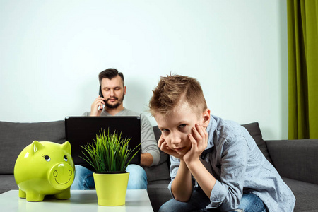 一个悲伤的男孩看着相机，而他的父亲在家里的背景下工作。 时间的概念在一起，孤独的孩子在家里工作，自由职业者复制空间。