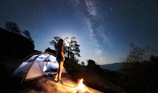 年轻女子徒步旅行者在篝火旁的岩石山顶上休息，夏季夜间还有发光的旅游帐篷