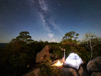 年轻女子徒步旅行者在篝火旁的岩石山顶上休息，夏季夜间还有发光的旅游帐篷