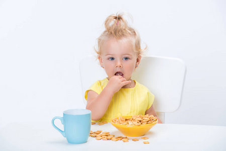 一岁的金发小女孩穿着黄色T恤，在白色背景的桌子上吃麦片粥，喝一杯奶茶。
