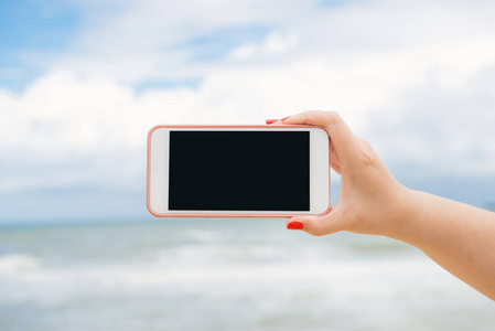 女人的手在沙滩上显示一个空白的智能手机横屏，背景是大海
