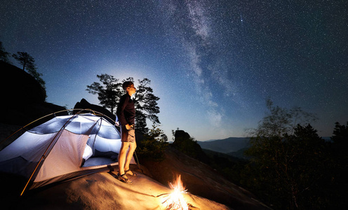 年轻的徒步旅行者在篝火旁的岩石山顶上休息，在夏季夜晚照亮旅游帐篷