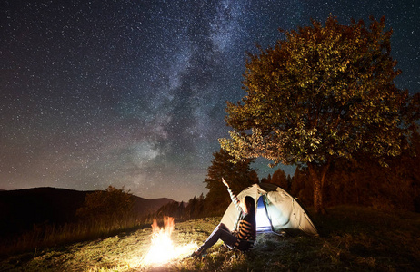 女游客休息在篝火旁露营，照亮旅游帐篷，欣赏充满星星和银河的夜空