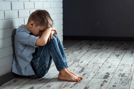 一个小男孩独自坐在墙附近的学校里，有一种悲伤的感觉。 被遗弃在走廊里的被冒犯的孩子弯着腰靠在砖墙上。 带有复制空间的欺凌歧视。