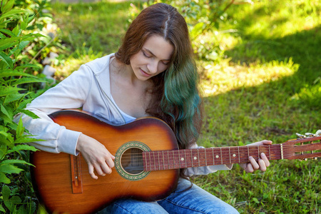 年轻的时髦女人坐在草地上，在公园或花园背景上弹吉他。 青少年女孩学习演奏歌曲和写作音乐。 爱好生活方式放松工具休闲教育观念