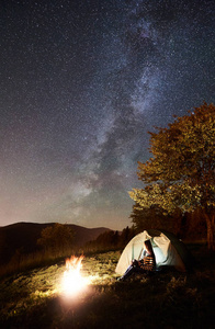 女游客休息在篝火旁露营，照亮旅游帐篷，欣赏充满星星和银河的夜空