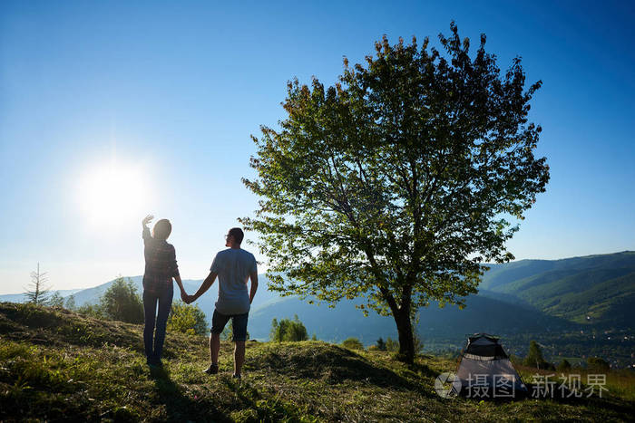 在阳光明媚的早晨,两个快乐的年轻人和女人徒步露营和大树附近休息