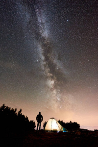 在山顶露营时，在发光的旅游帐篷旁休息的男性徒步旅行者的剪影