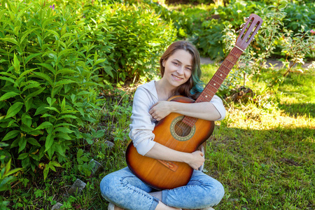 年轻的时髦女人坐在草地上，在公园或花园背景上弹吉他。 青少年女孩学习演奏歌曲和写作音乐。 爱好生活方式放松工具休闲教育观念