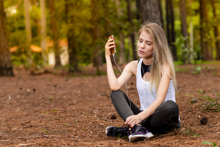 美丽的金发模特在松树中间坐着，用手机带耳机。 听音乐