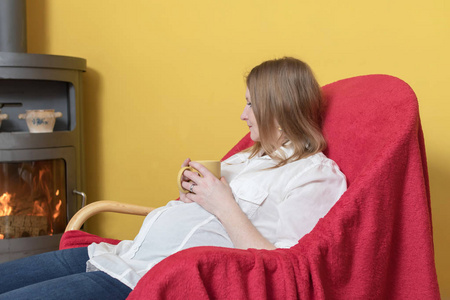 怀孕的年轻女子正在家中的壁炉炉子上坐在椅子上放松。 女人拿着黄色的杯子