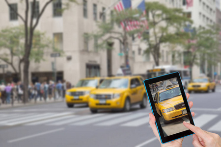 女性手指触摸平板电脑与黄色出租车的照片在触摸屏。 有意模糊的第五大道NYC的图像在背景中。 所有潜在商标都被删除。