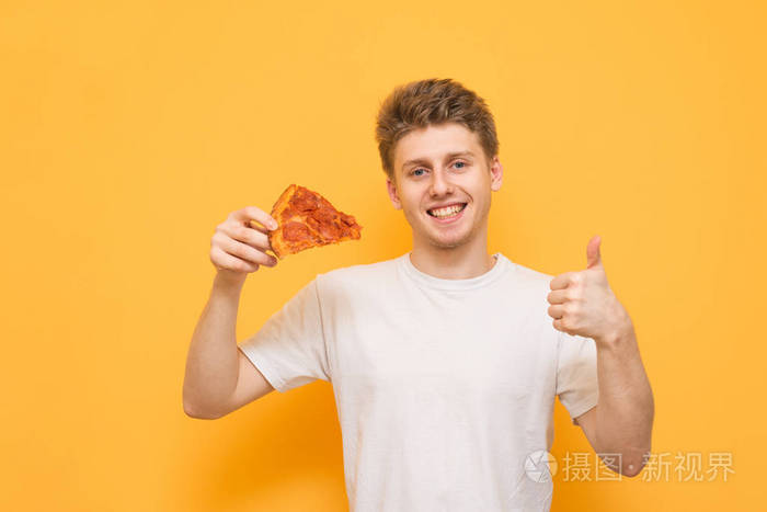 快乐的年轻人穿着白色T恤，手里拿着一块开胃的披萨，看着相机，拇指向上。 微笑的家伙手里拿着披萨，在黄色的背景上被孤立了。