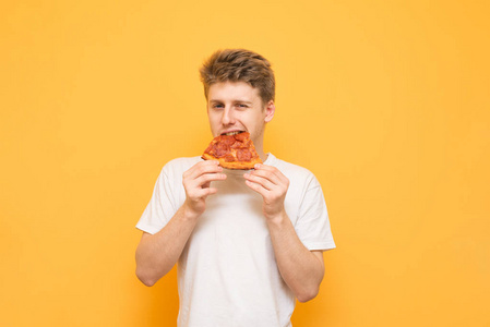 英俊的年轻人咬了一块开胃的披萨，看着黄色背景上的相机。 穿白色T恤的男孩吃黄色背景下孤立的快餐披萨。 共用空间