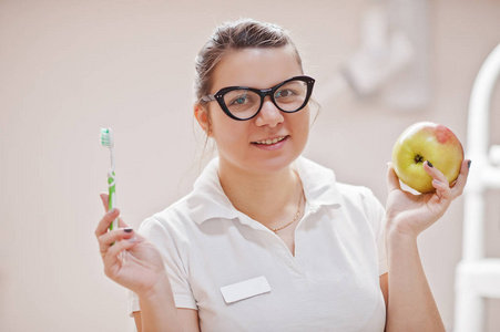 特写戴眼镜的女牙医的肖像，她站在牙科办公室，手里拿着一个苹果和牙刷。