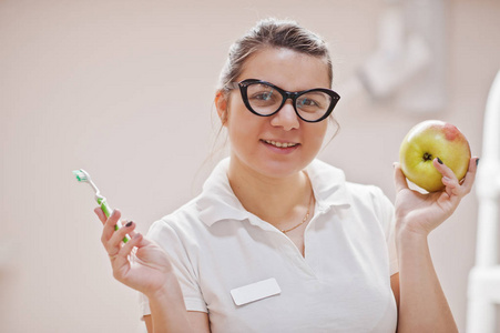 特写戴眼镜的女牙医的肖像，她站在牙科办公室，手里拿着一个苹果和牙刷。