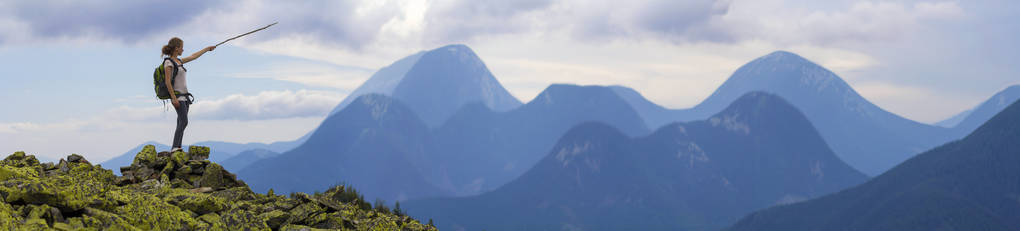 年轻的苗条金发旅游女孩与背包点与棍子在雾山山脉全景站在岩石顶部明亮的蓝色早晨的天空背景。 旅游旅游和攀岩概念。