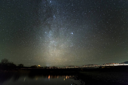 地平线上鹅卵石河岸树木的景观，明亮的星星和银河系在黑暗的天空中反射在平静的水中。 自然概念的美。