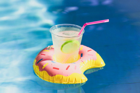 在游泳池里, 在充气甜甜圈玩具上, 品尝冰和新鲜的柠檬汁的美味鸡尾酒。夏季在游泳池度假和放松时间。带阳光倒影的游泳池里的波纹水