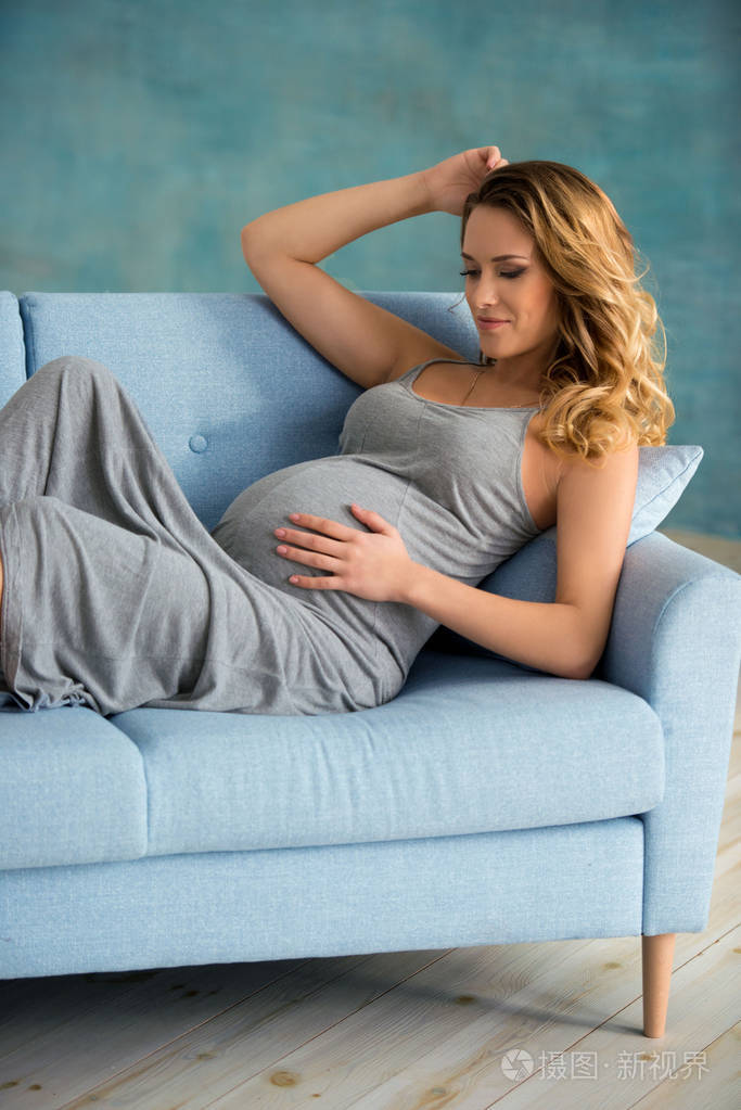 美丽快乐的年轻孕妇穿着一件灰色的长裙坐在家里的沙发上