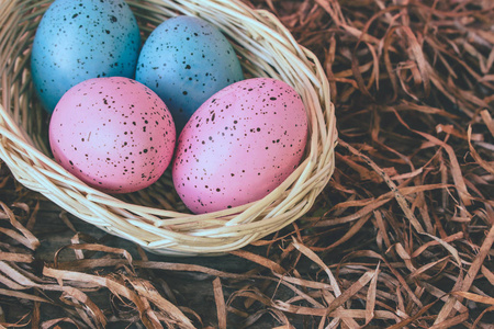 四个粉红色和蓝色复活节鸡蛋在一个篮子上的木制地板覆盖稻草背景。 复活节庆祝壁纸与复制空间。 古董加工。
