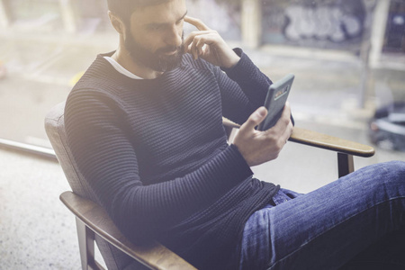 穿着黑色套头衫的头戴的头戴胡子的男子使用手机应用程序的手机博客。自信的年轻同事坐在现代阁楼的扶手椅上阅读社交网络上的新闻手机