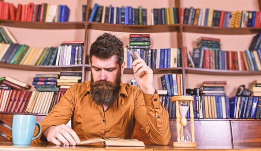 男人在忙碌的脸上看书, 书架上的背景。教育和科学概念。在图书馆学习胡子的老师或学生。科学家读书, 坐在桌与杯子和沙漏