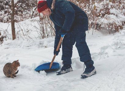 一个拿着铲子的人在降雪时从雪中清理道路。猫就在附近