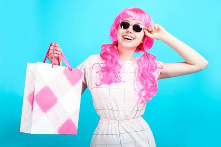 美丽的微笑女孩，穿着鲜艳的粉红色头发，蓝色背景上有购物袋。 概念销售购物。
