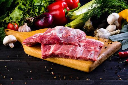 生猪肉。 新鲜牛排在木切割板上的深色背景。 带蔬菜和香料的生肋排胸