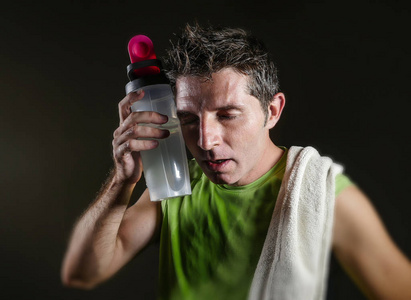 年轻的疲惫和疲惫的运动男子，肩膀上有毛巾，出汗的饮用水瓶，经过艰苦的锻炼和健身训练，在健康的生活方式概念中孤立于黑暗的背景中