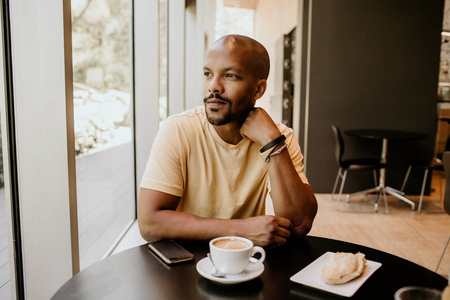 年轻的英俊的非洲男人坐在现代城市的咖啡旁, 望着窗户