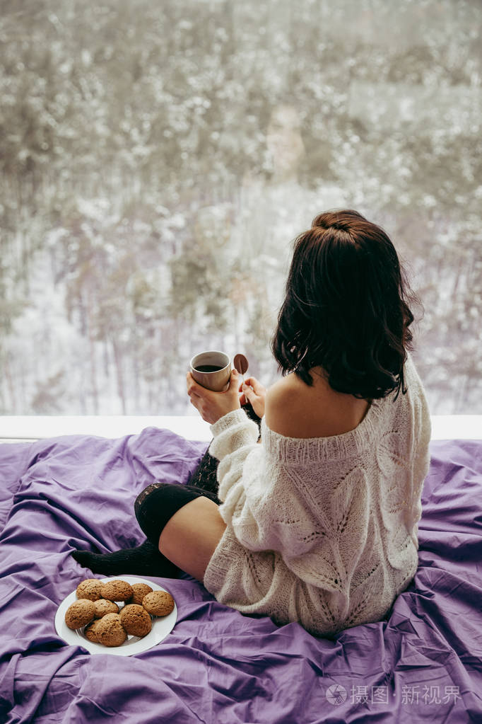 年轻的黑发女人坐在窗边喝着咖啡和饼干的茶