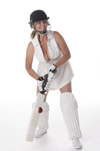 女板球运动员穿着白色连衣裙，戴着安全帽，小腿垫着蝙蝠和球。