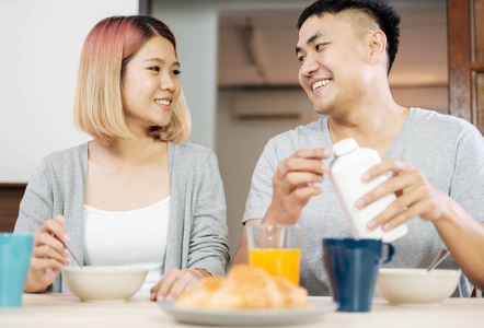 快乐的亚洲夫妇穿着睡衣，早上坐在厨房里，一起吃麦片早餐。丈夫把牛奶倒进妻子的碗里