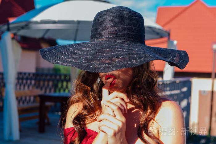 美丽的年轻女子，穿着红色的衣服，戴着帽子，在夏天阳光明媚的日子里，用手指捏着嘴唇