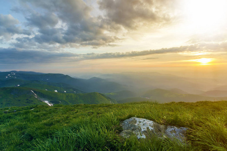 日出时广阔的夏季山景。 明亮的橙色太阳在蓝色多云的天空中升起，在绿色的草山上，大岩石和遥远的山脉覆盖着晨雾。 自然概念的美。