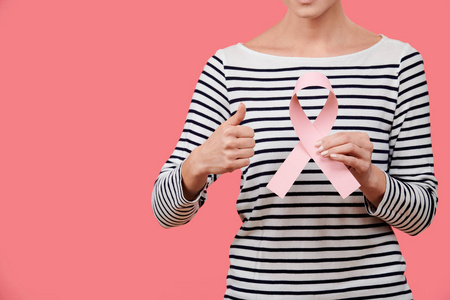 一位年轻女子手持粉红色乳腺癌意识丝带，竖起大拇指，被隔离在珊瑚背景上。医疗保健人慈善和医学概念。
