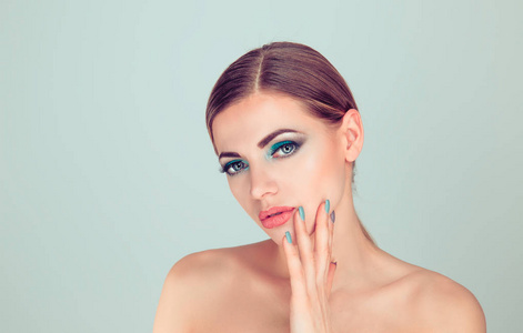 的女人用手摸着脸，用化妆做指甲。漂亮的年轻模特，蓝绿色的眼影化妆和指甲隔离在浅绿色灰色背景。