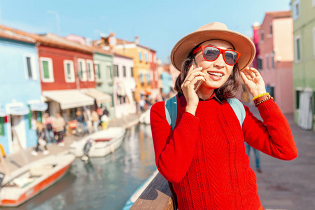 年轻快乐的旅行者女人在威尼斯布拉诺岛的五颜六色的房子里通过电话交谈。意大利旅游概念