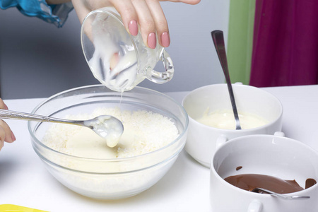 女人在椰子薯片中加入炼乳。 甜点的配料在桌子上。 用椰子和炼乳烹饪糖果。 在白色和黑色巧克力的釉里。