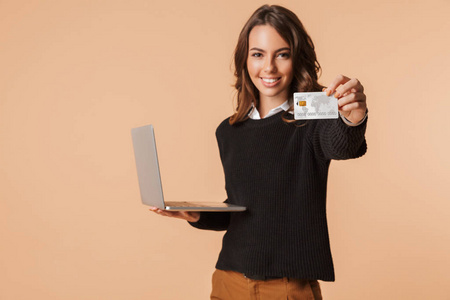 兴奋的年轻女子穿着毛衣，手提电脑显示塑料信用卡与米色背景隔离